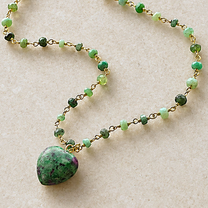 A beaded necklace with green and light green faceted beads on a gold-tone chain, featuring a heart-shaped green stone pendant, displayed on a light beige textured background. The brand logo 'THE SILVER WREN' is visible in the top left corner.