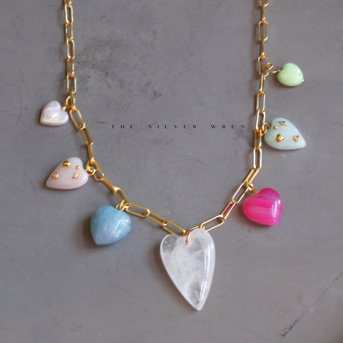 A gold-plated paperclip chain necklace with multiple quartz stone heart charms in various colors.