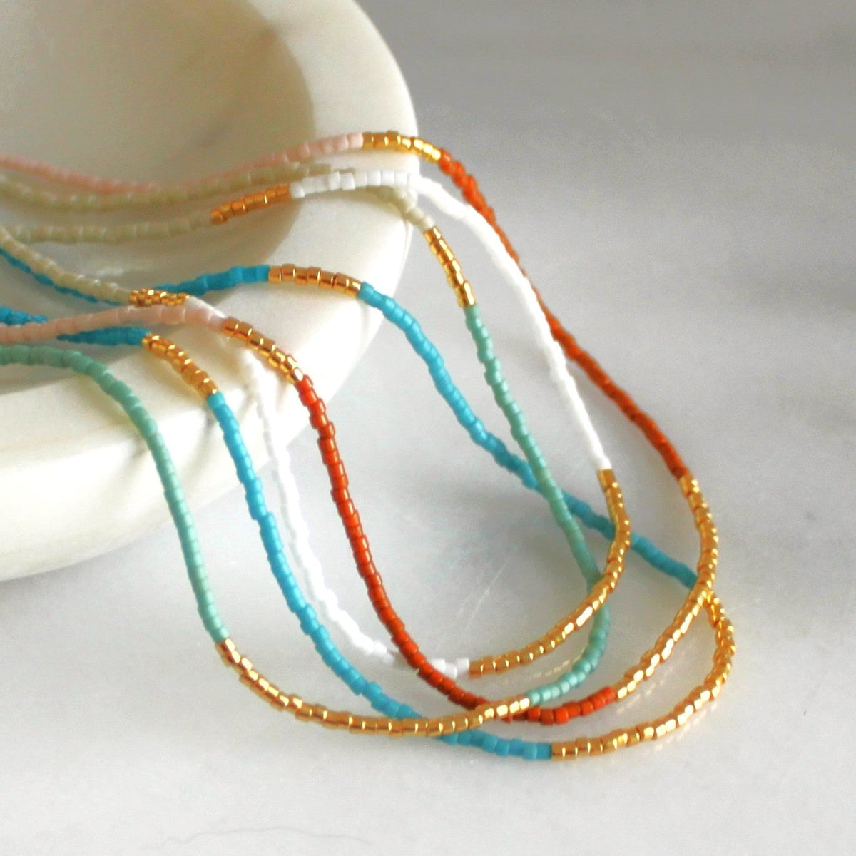 A close-up of delicate beaded strands draped over a white ceramic dish on a light marble surface. The beads are arranged in thin strands of white, turquoise blue, orange, and metallic gold.