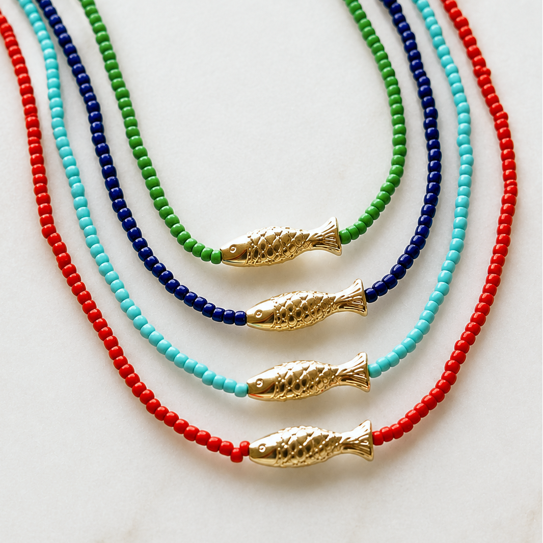 Four beaded necklaces arranged on a light marble surface, each featuring a small gold fish pendant. The necklaces use red, turquoise, light blue, and dark blue beads, with the gold fish pendants positioned near the bottom of each strand.