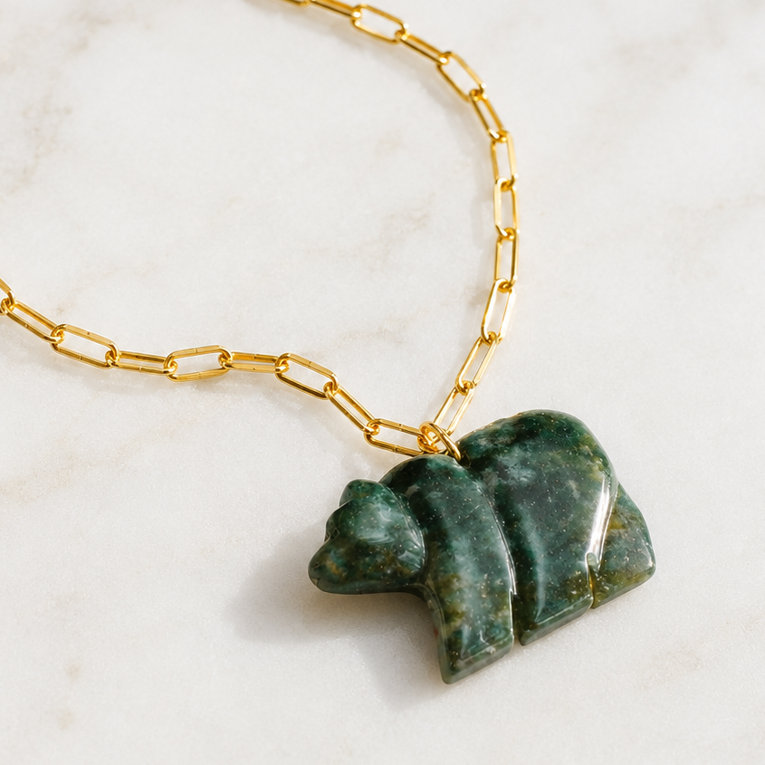 A gold chain necklace with a carved green stone bear pendant, displayed on a white marble surface.