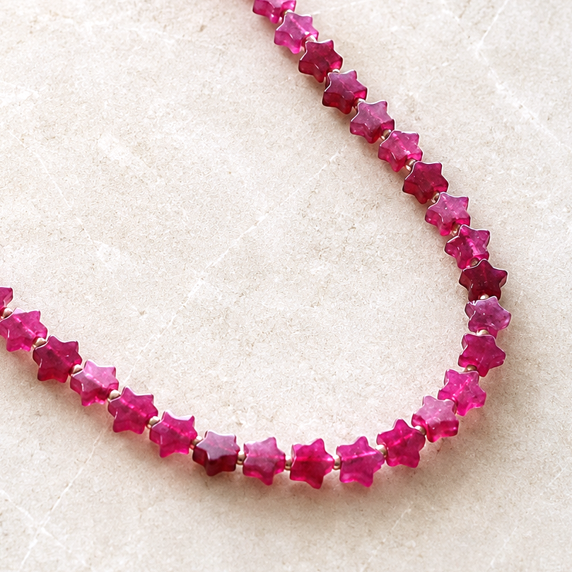 A strand of faceted star-shaped pink gemstones, likely rubies, arranged in a U-shape on a light beige textured surface. The brand logo 'THE SILVER WREN EST.
