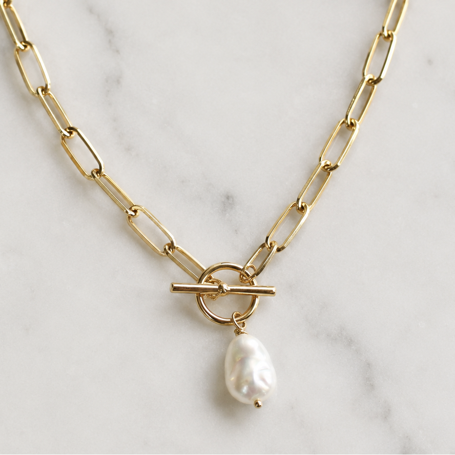 A gold paperclip chain necklace with a toggle clasp and a dangling baroque pearl, displayed on a white marble surface.