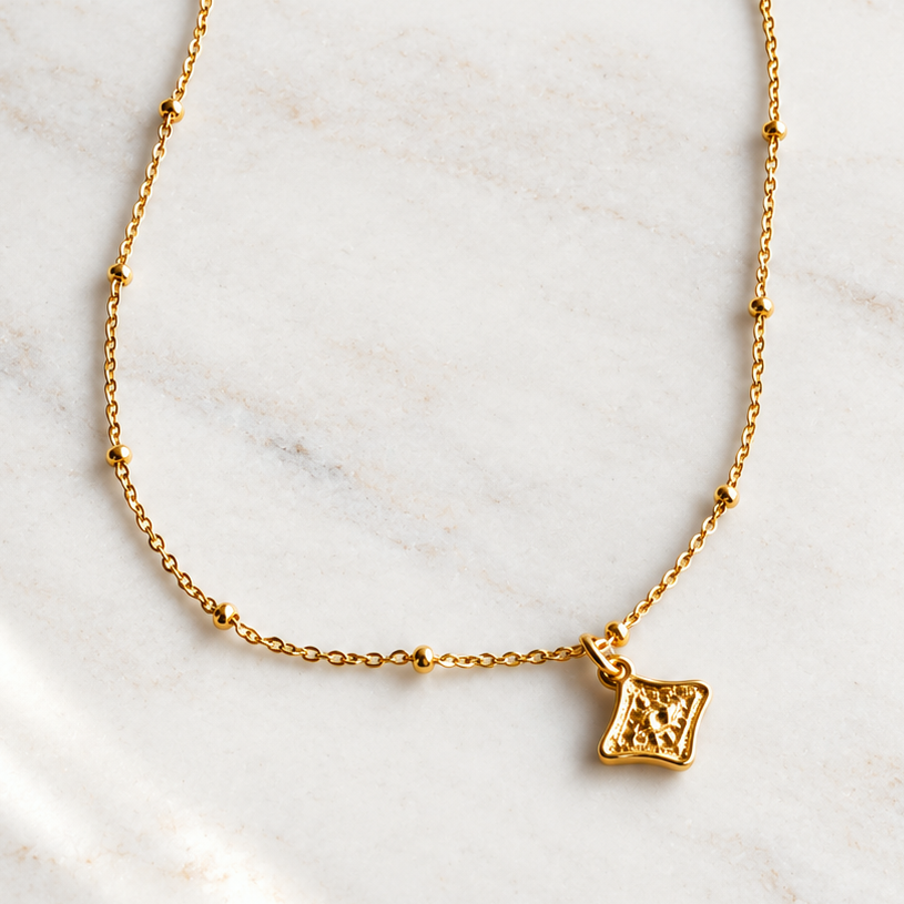 A delicate gold chain necklace with a small square pendant featuring an engraved cross, displayed on a light marble surface with soft shadows.