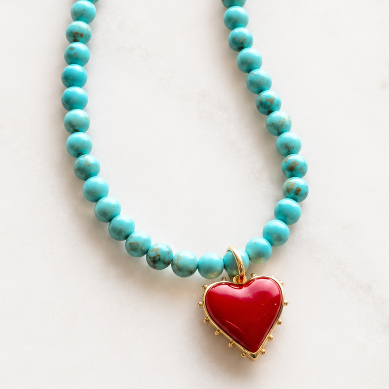 A turquoise beaded necklace with a red heart-shaped pendant on a light marble background. The brand logo 'The Silver Wren' is visible in the top left, alongside product feature icons.