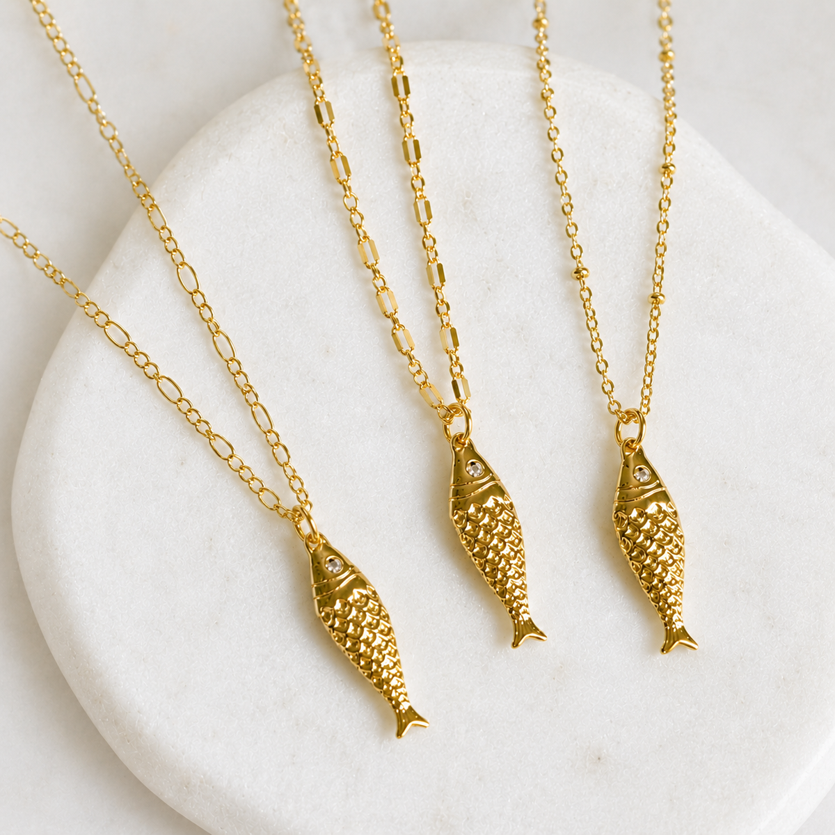 Three gold fish-shaped pendant necklaces with textured scales and small clear stones, displayed on a white stone surface.