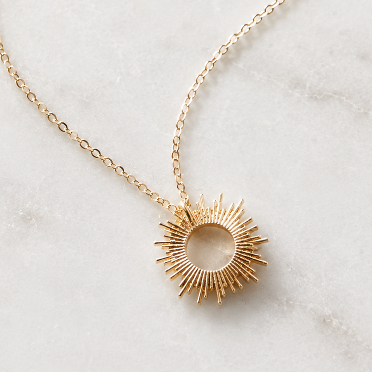 A gold necklace with a sunburst pendant is displayed on a light marble background. The brand name 'THE SILVER WREN' is printed at the top, and several feature icons and text are listed on the left side.