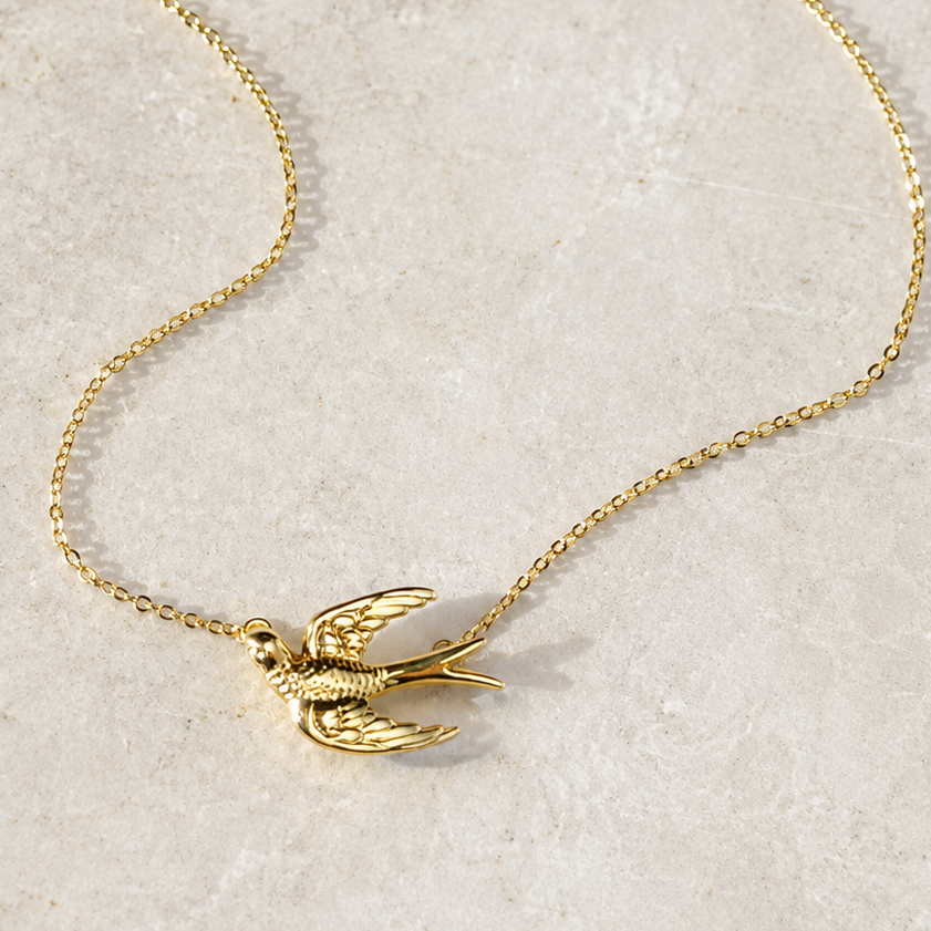 A delicate gold chain necklace featuring a small bird pendant with spread wings, displayed on a light beige textured stone surface.